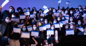 Foto de diversos recém-formados sobre um palco. Alguns estão sentados, enquanto outros estão atrás, de pé. São dezenas de jovens homens e mulheres, a maioria brancos. Todos usam máscaras, roupas em estilo formal ou esporte fino e posam para a câmera segurando ou agitando seus certificados de formatura.