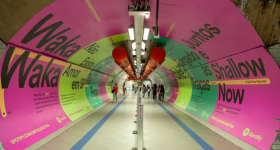 Foto tirada na ponta de um corredor no metrô de São Paulo . As paredes estão totalmente cobertas por cartazes publicitários chamativos (predominantemente em cor-de-rosa e em dois tons de verde) com títulos de músicas famosas escritas em preto. O corredor é bem iluminado. Nele, algumas barras de segurança separam as pessoas que andam nos dois sentidos.