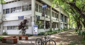 Foto do prédio do CTR. Em primeiro plano, há uma placa azul onde está escrito ECA em branco. No poste que sustenta a placa está amarrada uma bicicleta. Na parte esquerda existem três bancos laranja escuro, sem encosto. Mais ao fundo localiza-se o prédio, que foi fotografado de lado, de modo que é possível ver os dois lados dele. Na frente do prédio há placas em azul identificando o nome e a sigla do departamento. Ao seu lado encontra-se um caminho, ladeado de grama, arbustos e árvores. 