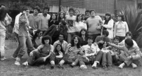Foto em preto e branco mostra diversos jovens, homens e mulheres, no gramado em frente à fachada do prédio principal da ECA. Alguns estão sentados, enquanto outros estão atrás, de pé. A maioria sorri posando para a câmera e abraçando os colegas, enquanto outros estão distraídos no momento da foto. Alguns ainda fazem brincadeiras com os colegas. 