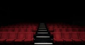 Foto de sala de cinema vazia. Ao centro, há um corredor de escadas. Várias fileiras de poltronas vermelhas podem ser vistas, todas elas vazias. O fundo da imagem é escuro, uma sombra.