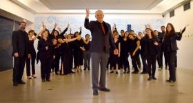 Foto dos integrantes do Coro de Câmara Comunicantus. Eles estão em uma sala. À frente do grupo, está regente e professor do coro. Ele é um homem mais velho, com cabelos curtos e brancos nas laterais da cabeça, usa um terno preto com gravata vermelha e está com uma das mãos levantadas. Ao fundo, está o grupo. Todos usam roupas pretas e olham para a câmera, com os braços levantados.