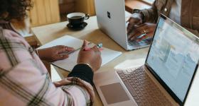 Imagem de duas pessoas trabalhando em casa. Ambas possuem a pele preta e estão utilizando computadores, que estão em cima de uma mesa. Em primeiro plano, uma mulher com anéis nos dedos e caneta na mão escreve em um caderno. Em segundo plano, outra mulher digita. Ao fundo, mas ainda em cima da mesa, há uma xícara preta com café.