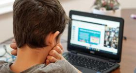 Foto de um menino sentado assistindo à aula online. Ele tem a pele branca, cabelos curtos e escuros, usa uma camiseta cinza e está com o rosto apoiado nas mãos semifechadas. O fundo é desfocado, mas é possível observar um computador sobre a mesa. O computador mostra uma apresentação de slides dividindo espaço com uma videochamada.