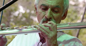 Foto de Toninho Carrasqueira, homem branco, idoso de cabelo brancos e bigode grisalho que usa uma camisa branca e toca uma flauta transversal. 