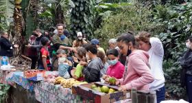 Foto de uma mesa ou bancada com toalhas coloridas estendidas. Em cima da mesa há várias frutas dispostas: mexericas, bananas, maçãs e melancias. Em volta da mesa há uma grande quantidade de pessoas em pé, entre crianças e adultos, algumas escolhendo frutas e outras mais afastadas.  Ao fundo há várias árvores com diversos tipos de folhagem.