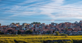 Foto de comunidade periférica próxima a um rio. Em primeiro plano, é possível observar parte do leito do rio e um campo de grama verde. Na parte superior, existem vários fios de alta tensão. Em segundo plano, há muitas casas próximas umas das outras, a maioria com tijolos e cimento aparentes. Existem poucas árvores no local. Ao fundo, um céu azul com algumas nuvens dispersas.