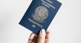 Foto. A frente de um fundo branco, uma mão, de uma pessoa branca, segura um passaporte, um livreto azul escuro com informações em dourado.