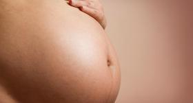 Foto de uma grávida. Apenas sua barriga, grande por conta da gravidez, aparece na imagem. A mão da mulher, que é branca, está apoiada em cima da barriga. O fundo da imagem é totalmente rosa.