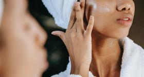 Foto de uma mulher negra com uma toalha no cabelo passando creme em frente ao espelho. No canto esquerdo da imagem, aparece o seu rosto desfocado. Em segundo plano e em foco, a mulher passa o creme em sua bochecha. É possível ver apenas seu nariz e sua boca. Ela veste um roupão de banho branco. 