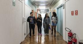Foto de quatro estudantes andando em um corredor. À esquerda, um menino e uma menina estão conversando, eles são brancos e vestem roupas pretas. Ao lado deles, duas jovens se olham e sorriem, a da esquerda é branca e veste uma roupa preta com bolinhas pretas, a da direita é negra e veste cinza. No corredor, há algumas portas laranjas e azuis, as paredes são brancas.