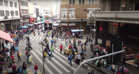 Print de um trecho do documentário 25 de Março: a singularidade dos fluxos e fixos. A imagem mostra, a partir de um ângulo elevado, as esquinas da rua 25 de março com a rua Cavalheiro Basílio Jafet e a Ladeira Porto Geral. Muitas pessoas estão nas calçadas e no meio da rua, vendendo ou comprando. 