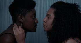Foto de uma cena do curta. Duas mulheres negras se olham profundamente nos olhos. A da esquerda usa um brinco e tem cabelo crespo e curto. A da direita tem cabelo cacheado e volumoso. Ela coloca sua mão esquerda no pescoço da outra mulher. 