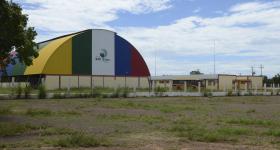 Foto da escola do quilombo Mata Cavalo tirada de longe. Em mais evidência, está a quadra da escola, que possui uma cobertura semicircular colorida. Mais ao fundo, está o prédio das salas de aula. O prédio é comprido e possui paredes amarelas, com listras vermelhas e muitas janelas. Na frente da construção, há um ônibus escolar amarelo. O chão é de terra e há nele muitas pedras, além de alguns pontos com grama.