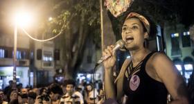 Foto de Marielle Franco, uma mulher negra que usa uma regata preta, brincos redondos pretos e uma faixa colorida no cabelo. Ela fala em um microfone. Há um adesivo lilás no seu peito e um livreto roxo na outra mão, ambos com seu nome escrito. Ao fundo, pessoas ouvem a vereadora. Entre essas pessoas, há dois fotógrafos, que apontam câmeras para ela.