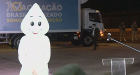 Foto do personagem Zé Gotinha sozinho na pista do aeroporto. Ele está ao lado de um pequeno púlpito de acrílico e ao fundo está estacionado um caminhão azul da campanha de vacinação. Na parte direita da foto, alguns soldados do exército armados se posicionam embaixo de um grande avião. 