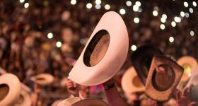 chapeu de cauboi é erguido por uma mãos de um homem branco, em meio a uma multidão de um show. ao redor, outros chapeus aparecem ergidos; ao fundo, luzes desfocadas