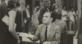 Foto em preto e branco. Ao centro, Nelson Rodrigues está sentado em uma cadeira, a frente de um espelho e atrás de uma mesa de madeira. É um homem branco de meia idade, de cabelo grisalho e usa um terno. Segura uma caneta com a mão esquerda e com a direita estende um livro a uma mulher, que usa um vestido preto, é branca e tem cabelo claro e curto. No reflexo do espelho ao fundo há uma fila de pessoas, algumas com um livro na mão.