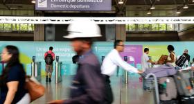 Foto do vai e vem de transeuntes com bagagens no terminal de passageiros do Galeão. Ao fundo, placa informativa  com o ícone de um avião subindo e os dizeres "Embarque doméstico" em português e inglês. 