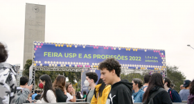 Foto de diversas pessoas caminhando entre as grades que organizam a fila de entrada para a Feira USP e As Profissões. Ao fundo, uma estrutura com o nome do evento e os dizeres "Boas Vindas". Atrás dessa estrutura está a Torre do Relógio.