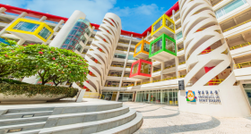 Foto da fachada da universidade. O prédio apresenta uma estrutura moderna, com três torres brancas espiraladas e alguns cubos coloridos projetados para fora da fachada. No canto inferior esquerdo da imagem, encontra-se uma parte elevada, na qual há um canteiro grande e florido.  