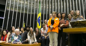 Foto de diversas mulheres, de diferentes idades e de maioria branca, na mesa e no púlpito da Câmara dos Deputados. Elas estão em pé, mas algumas estão mais altas do que outras devido ao desnível entre a mesa e o púlpito. Atrás das parlamentares está hasteada uma bandeira do Brasil.