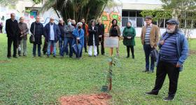Foto de um grupo de pessoas em um gramado. Em primeiro plano, uma muda de árvore que acaba de ser plantada ao lado de uma placa em memória de Ciro Marcondes. Ao lado, um homem negro de barba grisalha e dreadlocks na altura do ombro fala. Ao fundo, homens e mulheres de diferentes idades observam a cena atentos. 