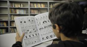 Foto de um menino branco, com cabelos curtos e castanhos que está sentado de costas. Ele lê um livro de histórias em quadrinhos. Ao fundo, estantes de livros.