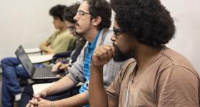 Foto de estudantes durante aula. Em primeiro plano, um homem negro de barba e cabelos escuros e crespos em penteado black power e óculos tem uma das mãos encostada no queixo. Atrás dele estão outros três jovens. Todos estão sentados em carteiras.