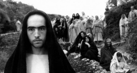 Foto em preto e branco. À esquerda, um homem branco usa uma veste branca e, por cima de sua cabeça e caindo sobre seu corpo, um tecido escuro. Ao fundo, há outras pessoas, que usam roupas semelhantes.