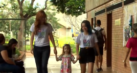 Foto de duas jovens caminhando de mãos dadas a uma menina. A menina tem cabelos castanho claros, usa vestido estampado e está no meio das moças, que usam uma camiseta branca com o logotipo colorido do projeto VOA. Ao fundo, calçadas e a fachada de um prédio da Universidade de Brasília.
