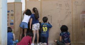 Foto de seis crianças desenhando e pintando em um mural de papel colado nas paredes e no chão. Algumas delas estão sentadas e outras estão em pé. 