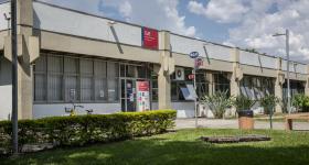 Foto da fachada do Departamento de Jornalismo e Editoração. Em primeiro plano, há um gramado ladeado por alguns arbustos. Mais ao fundo, está o prédio, onde encontram-se as placas do CJE e uma outra da ECA, que indica as direções para os outros departamentos. 