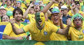Foto de torcedores do Brasil em um estádio de futebol. Em primeiro plano, aparecem quatro torcedores, com destaque para um homem branco, vestido com a camisa da Seleção, que está com cada lado do rosto pintado de verde e amarelo e com os braços erguidos. Um outro torcedor usa um chapéu com as cores da bandeira, e um terceiro, uma peruca e óculos escuros. Todos os quatro olham para a câmera. 