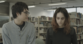 Imagem do filme Diário de Viagem. Um rapaz oriental de óculos observa menina de cabelo liso e pele pálida e com olhar baixo. Ao fundo uma prateleira de livros indicam que estão em uma biblioteca 