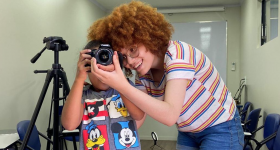 Foto de uma jovem ensinando uma criança a utilizar uma câmera fotográfica digital. A jovem tem pele clara, cabelos crespos castanho claros e óculos de aro redondo. O menino tem pele negra e cabelos curtos e raspados. Ele segura a câmera com o visor na frente de seus olhos, enquanto a jovem manuseia o anel de foco da lente junto com a criança.