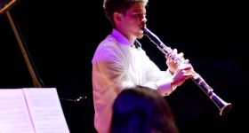 Foto de um homem tocando clarinete. Ele é branco, tem cabelos curtos e escuros e veste uma camisa branca. O fundo da imagem é totalmente preto, e o jovem é iluminado por luzes branca e roxa. Na porção inferior da foto, aparecem a cabeça de uma mulher que está de costas para a câmera e uma partitura apoiada no piano que ela toca.