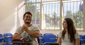 Foto de duas pessoas sentadas em uma sala de aula. Um homem sorridente está gesticulando com as mãos à esquerda. Ele é jovem, branco, tem óculos e cabelos escuros e veste uma camiseta clara com personagens desenhados. Uma mulher está sentada ao lado, ela olha para ele e sorri. A moça é jovem, é branca, tem longos cabelos lisos e escuros e veste uma camiseta branca. Atrás deles estão algumas carteiras escolares e uma janela.
