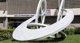 Foto de obra de arte sobre o gramado em frente à fachada do prédio principal da ECA. A escultura é branca, formada por 3 círculos e um semicírculo entrelaçados. Ao fundo, é possível ver a porta de entrada do prédio, com a inscrição Escola de Comunicações e Artes acima dela.