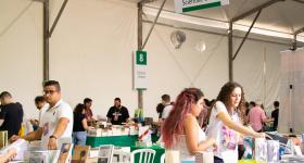 Foto do estande da editora Com-Arte na Festa do Livro da USP.  No expositor, os livros estão dispostos em uma mesa coberta por um tecido verde. Atrás dos livros, duas estudantes responsáveis pelo estade organizam as obras. Ao fundo, aparecem outros expositores do evento