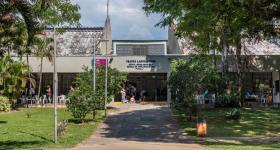 Foto da rampa de entrada para o prédio do Teatro Laboratório da ECA. As laterais da rampa têm um gramado arborizado. Ao fundo, bem no centro da imagem, a fachada do teatro. Há pessoas na entrada do prédio e cadeiras de plástico nas duas laterais da entrada. A luz do sol no chão indica que é um dia ensolarado.
