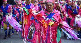 Foto de um senhor que veste um traje rosa de folia de reis e nas mãos usa uma bandeira com a imagem bordada de um santo e outros adornos. Ao fundo, foliões nas cores rosa e azul.
