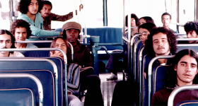 Foto de Milton Nascimento e do Clube da Esquina dentro de um ônibus antigo. São 13 homens ao total, 7 à direita e 6 à esquerda. Os bancos do ônibus são azuis e ele possui grandes janelas, que iluminam o interior do ambiente. 