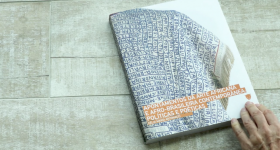Foto de um livro fechado sobre uma superfície de madeira clara. A capa do livro tem fundo branco e um tecido branco com palavras escritas em azul. Na porção inferior, estão o nome do livro e da autora e o logo da editora em branco sobre um fundo laranja. Uma mão de uma pessoa branca está apoiada na ponta do livro.