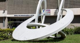 Foto de obra de arte sobre o gramado em frente à fachada do prédio principal da ECA. A escultura é branca, formada por 3 círculos e um semicírculo entrelaçados. Ao fundo, é possível ver a porta de entrada do prédio, com a inscrição Escola de Comunicações e Artes acima dela.