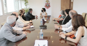 Foto de pessoas reunidas em uma grande mesa retangular. Na cabeceira da mesa, a cantora Marisa Monte - uma mulher branca, de cabelos escuros e ondulados, presos. Ela veste uma blusa branca e olha para a esquerda, onde está sentado o reitor da USP, um homem branco, de cabelos grisalhos e óculos. Ele veste um paletó cinza e uma camisa branca. Outras pessoas estão sentadas nos dois lados da mesa, homens e mulheres brancos. Ao fundo uma parede branca com o brasão da USP.