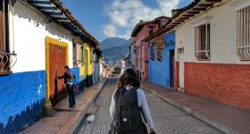 Foto de uma jovem que caminha sozinha por uma rua de Bogotá, na Colômbia. Ela está de costas para a câmera e carrega uma mochila preta nas costas. Seu cabelo é castanho e liso. Ela usa uma blusa cinza, de manga comprida, uma calça jeans e tem um casaco amarrado na cintura. A rua por onde ela passa é feita de tijolos e tanto do lado direito, quanto do lado esquerdo, existem casas em estilo colonial, coloridas nas cores branco, azul, rosa, verde, vermelho e amarelo.