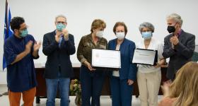 Foto de seis pessoas, três homens e três mulheres, em pé. Enquanto as mulheres seguram uma placa e um certificado, os homens aplaudem. Todos são brancos e usam máscara. Ao fundo, estão uma parede branca, uma mesa de madeira e uma bandeira azul hasteada. 