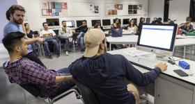 Foto de jovens do curso de Publicidade e Propaganda. São 20 estudantes sentados em uma sala, com cadeiras dispostas em círculo, entre homens e mulheres. Em primeiro plano, três jovens aparevem em frente a um computador, dois deles sentados e um de pé. A sala tem paredes brancas, com dois quadros com cartazes coloridos. Atrás dos estudantes sentados, em segundo plano, há dez computadores.