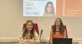 Foto de um seminário. Duas mulheres estão sentadas em uma mesa grande.  à esquerda, uma mulher branca, de cabelos longos e castanho claros. Ela tem um óculos apoiado sobre a testa. Veste uma blusa estampada e tem os antebraços apoiado um sobre o outro. à direita, uma mulher branca, de cabelos longos e castanho claros. Ela veste uma blusa laranja e um colar de madeira, marrom. à frente dela, um notebook aberto. Ao fundo, é projetada uma imagem da palestrante, título da palestra e outras informações.